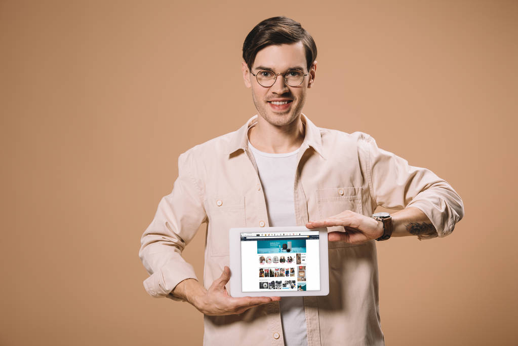 όμορφος άνδρας σε ποτήρια κρατώντας ψηφιακή δισκίο με amazon app στην οθόνη που απομονώνονται σε μπεζ  - Φωτογραφία, εικόνα