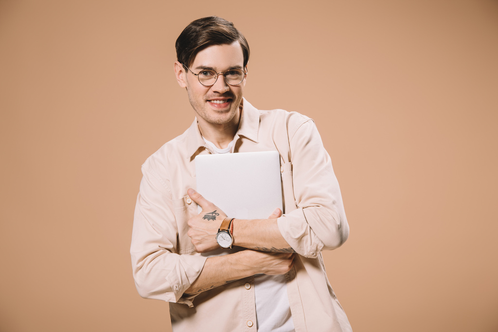 uomo allegro in occhiali che tiene il computer portatile isolato su beige
  - Foto, immagini