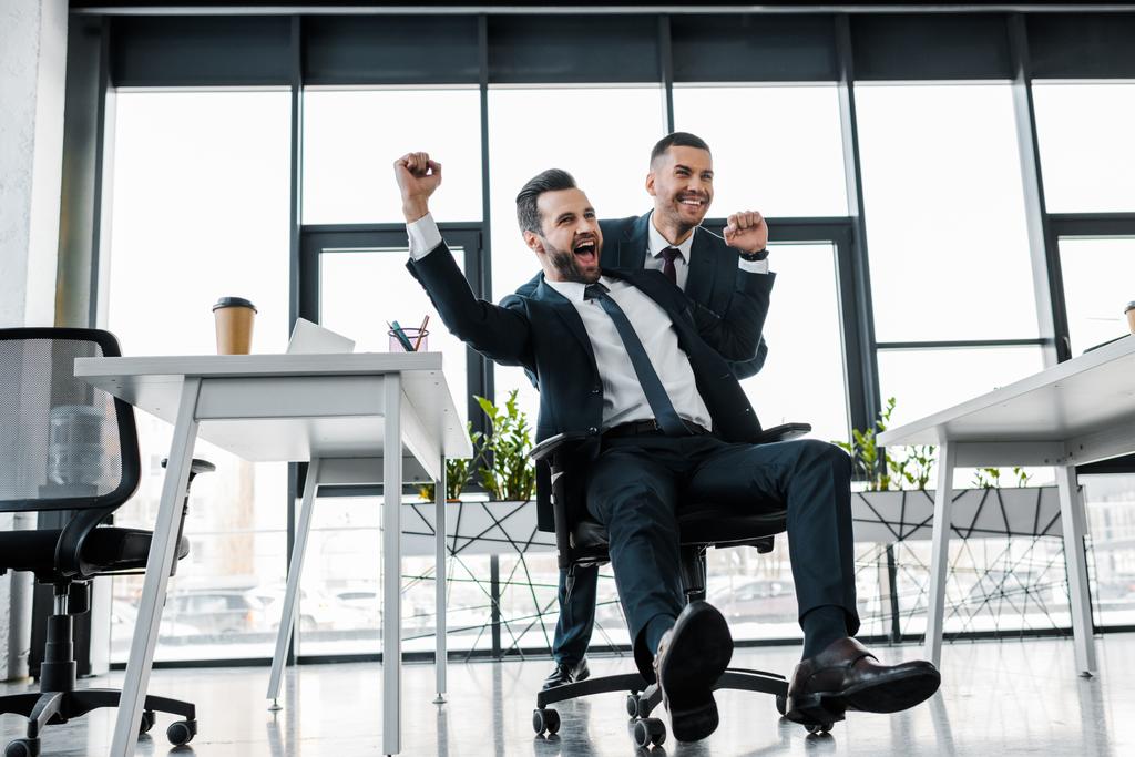 glücklicher Geschäftsmann reitet auf Stuhl neben Mitarbeiter in modernem Büro  - Foto, Bild
