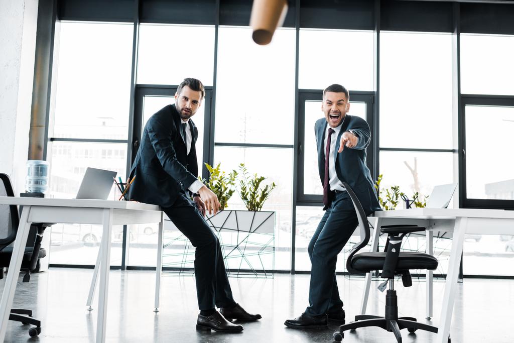 fröhlicher Geschäftsmann in formeller Kleidung zeigt mit dem Finger auf fallenden Pappbecher in modernem Büro  - Foto, Bild