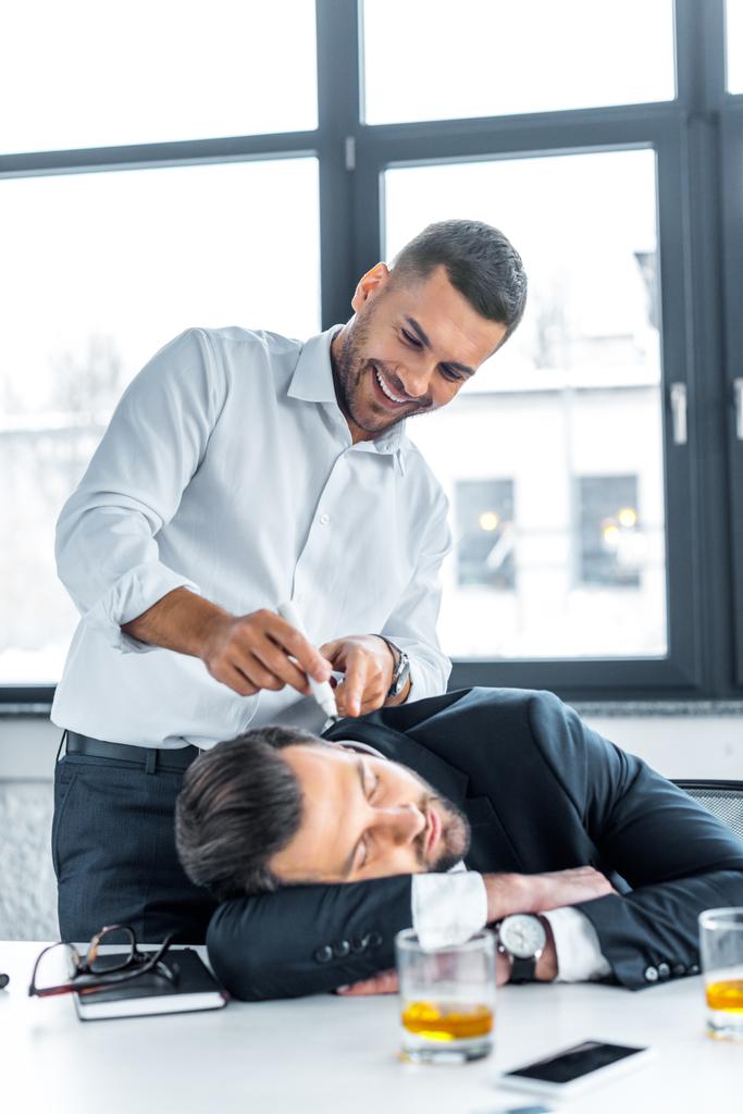 веселий чоловік, вказуючи з маркером на обличчі сплячого колеги в сучасному офісі
  - Фото, зображення