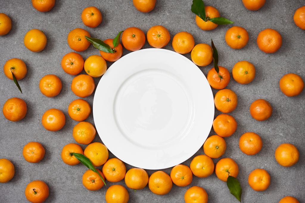 вид свежих органических мандаринов рядом с белой тарелкой на сером столе
 - Фото, изображение
