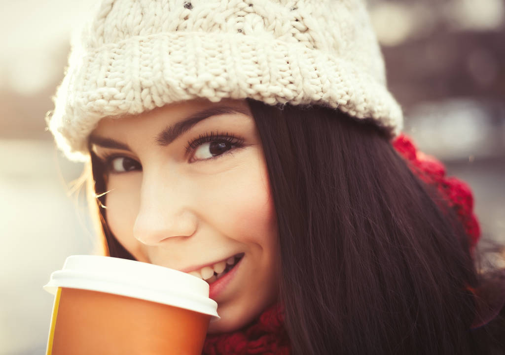 Μελαχρινή κορίτσι με ζεστά χειμερινά ρούχα πίνουν ζεστό καφέ υπαίθρια σε κρύα μέρα. Φωτεινό φωτισμένο πορτρέτο της ελκυστική ευτυχισμένη νεαρή γυναίκα - Φωτογραφία, εικόνα