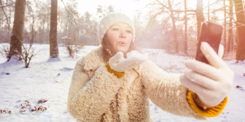 Szczęśliwa młoda kobieta dmuchanie pocałunek i biorąc selfie z smartphone w zimowym lesie - Zdjęcie, obraz