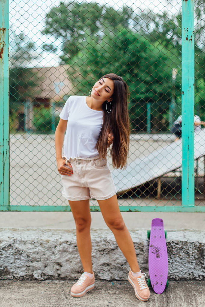 Летний образ жизни модной красивой молодой девушки, стоящей рядом с коартом скейтборда со своим фиолетовым пластиковым скейтбордом
. - Фото, изображение