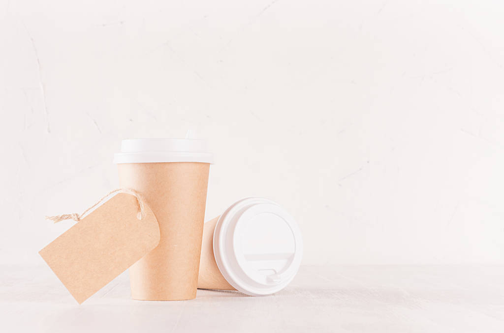 Coffee packing mockup - two craft brown paper cups, blank white caps and blank label on white wood board with copy space, coffee shop interior. Modern elegant concept for branding identity, advertising, design. - Photo, Image