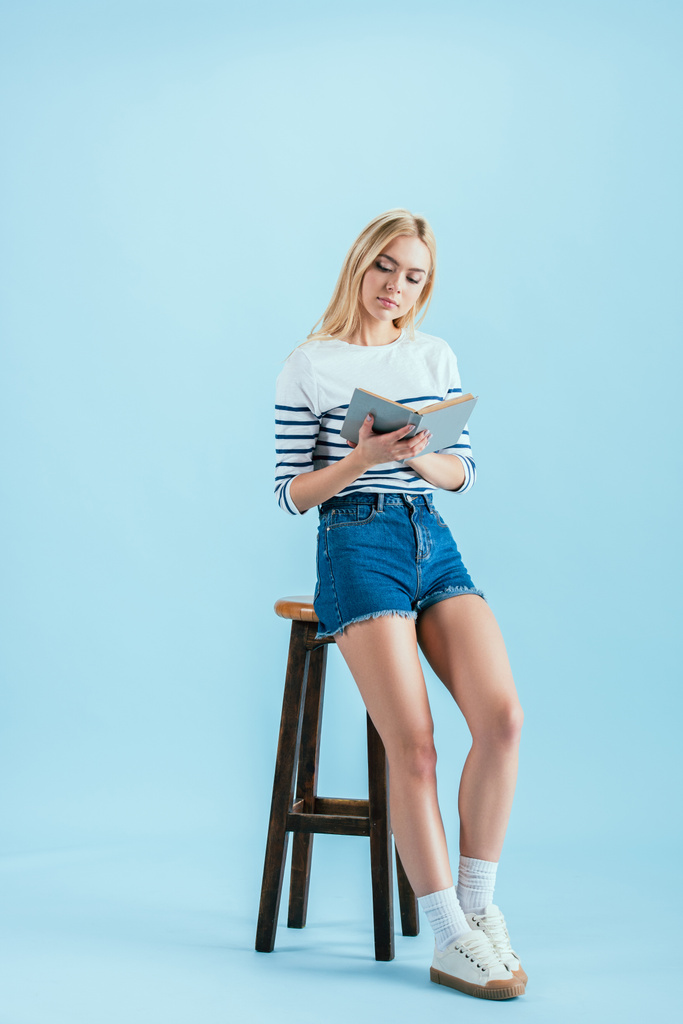 wunderbare junge Frau sitzt auf Stuhl und liest Buch auf blauem Hintergrund - Foto, Bild