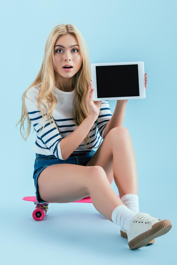 Chica rubia con el pelo largo sentado en el longboard y la celebración de la tableta digital con pantalla en blanco sobre fondo azul
 - Foto, Imagen