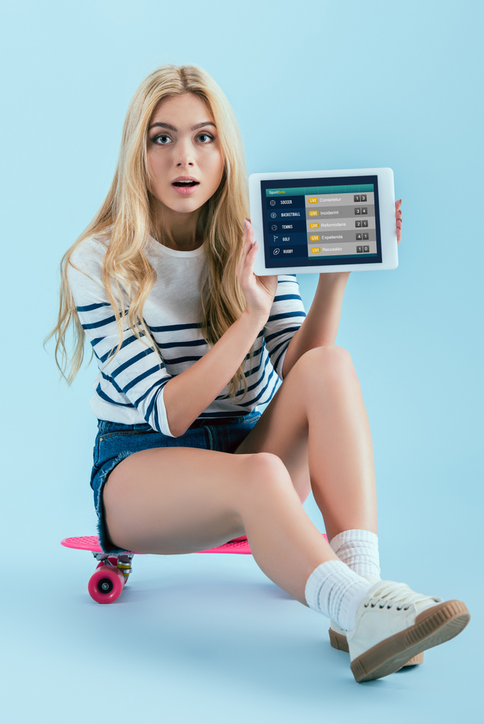 Menina bonita incrível sentado no longboard e segurando tablet digital com aplicativo sportbets na tela no fundo azul
 - Foto, Imagem