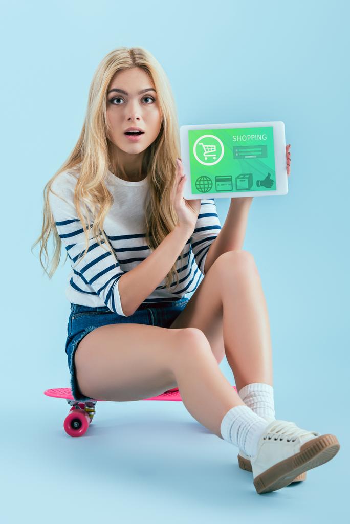 Шокована дівчина в джинсових шортах, що сидить на дошці і тримає цифровий планшет з онлайн-додатком для покупок на екрані на синьому фоні
 - Фото, зображення