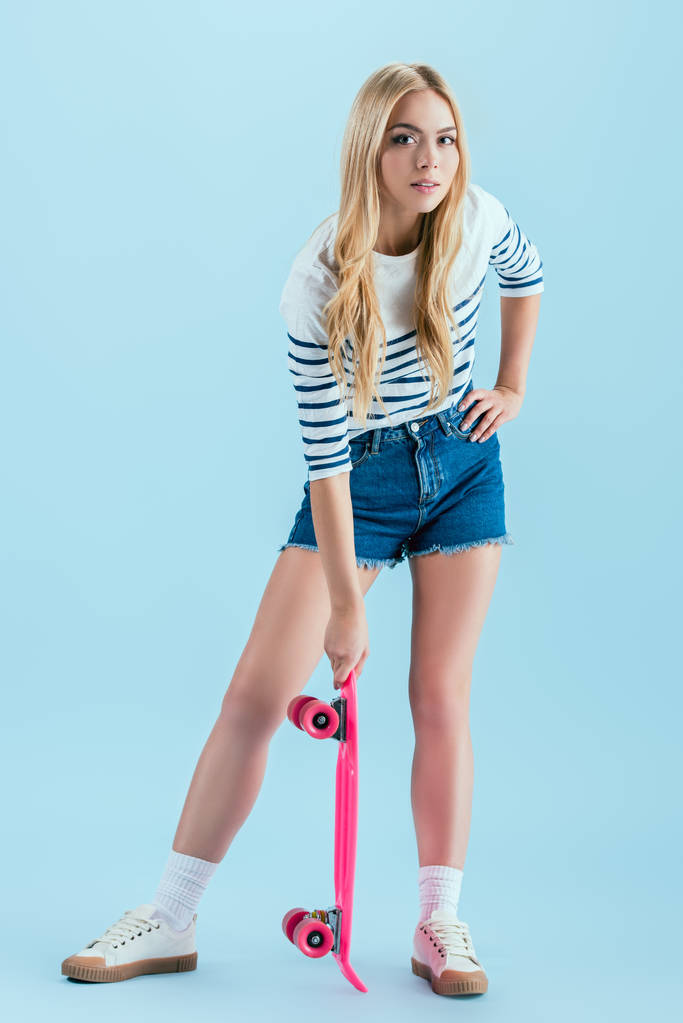 Charming blonde girl posing with pink longboard isolated on blue - Photo, Image
