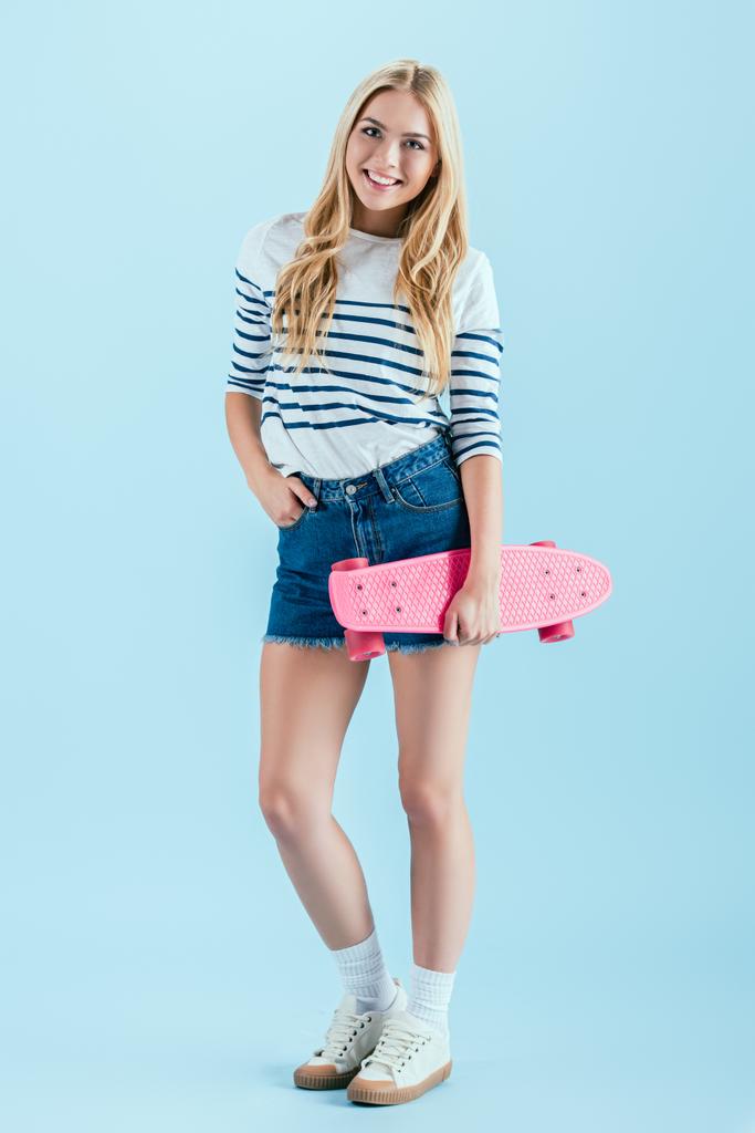 Opgewekt meisje met longboard poseren met de hand in de zak op blauwe achtergrond - Foto, afbeelding
