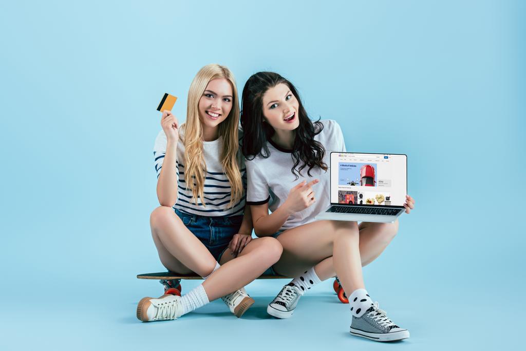 Estudio de fotos de las niñas que sostienen el ordenador portátil con el sitio web de ebay en la pantalla y la tarjeta de crédito mientras está sentado en longboard sobre fondo azul
 - Foto, Imagen