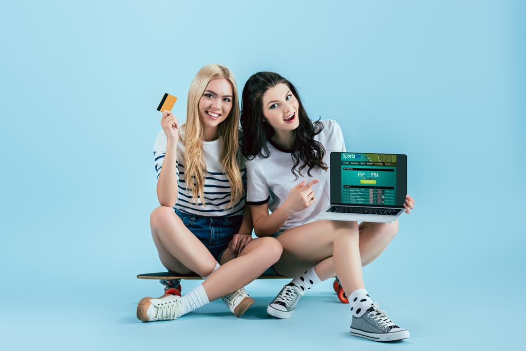 Viehättävä naiset tilalla kannettava sportsbet verkkosivuilla ruudulla ja luottokortilla istuu longboard sinisellä pohjalla
 - Valokuva, kuva