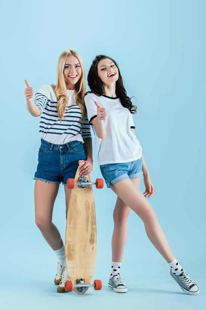 stylische Mädchen mit hölzernem Longboard posieren mit erhobenen Daumen auf blauem Hintergrund - Foto, Bild