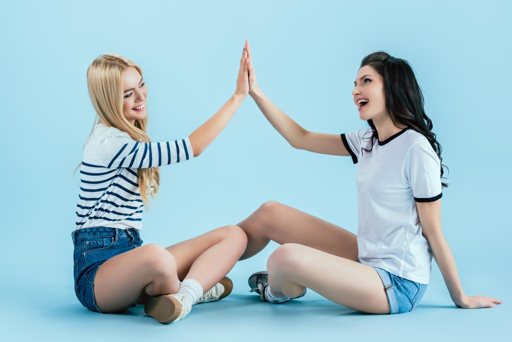 Cheerful girls in denim shorts posing on floor on blue background - Photo, Image