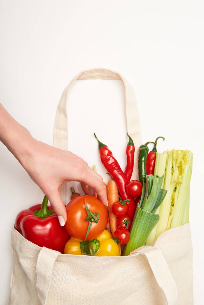 Vista parcial de la mujer sacando tomate de la bolsa con verduras sobre fondo blanco
 - Foto, Imagen