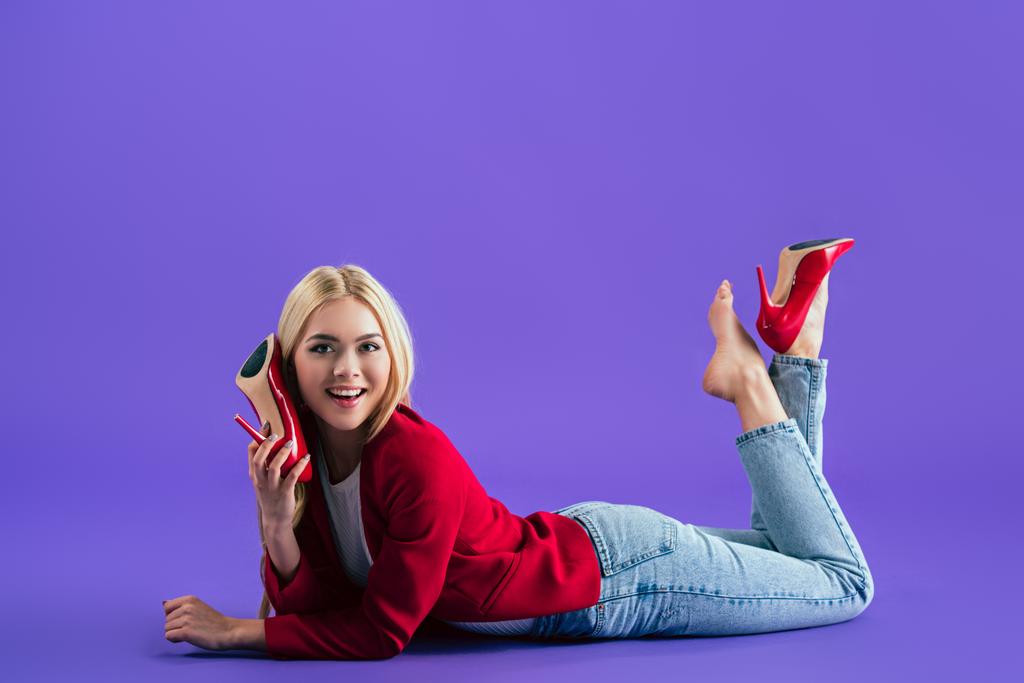 Чудова усміхнена дівчина в червоному взутті на високих підборах лежить на підлозі і дивиться на камеру на фіолетовому фоні
 - Фото, зображення