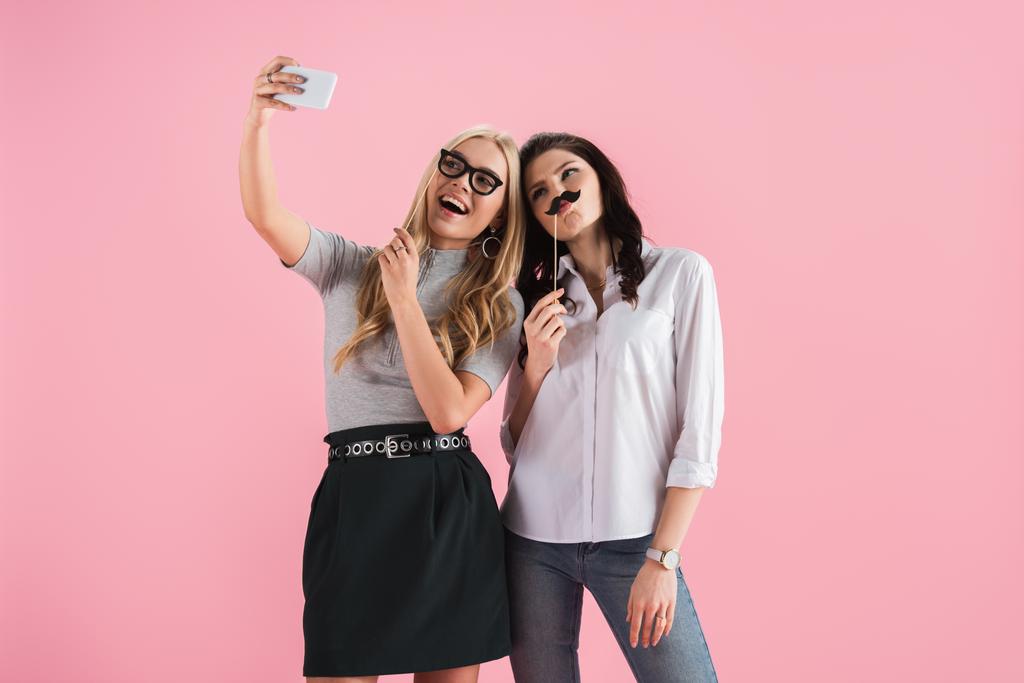 Meninas com bigode falso e óculos tomando selfie isolado em rosa
 - Foto, Imagem