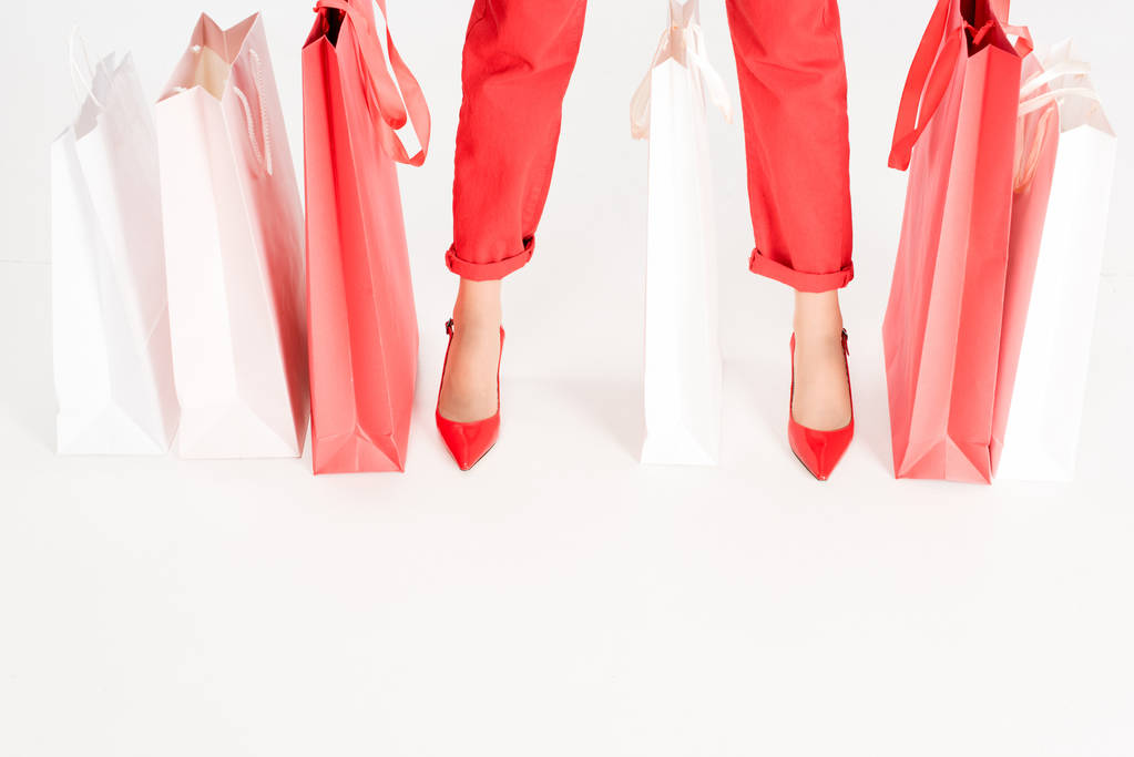 abgeschnittene Ansicht weiblicher Beine in der Nähe roter Einkaufstüten isoliert auf weißem Grund  - Foto, Bild