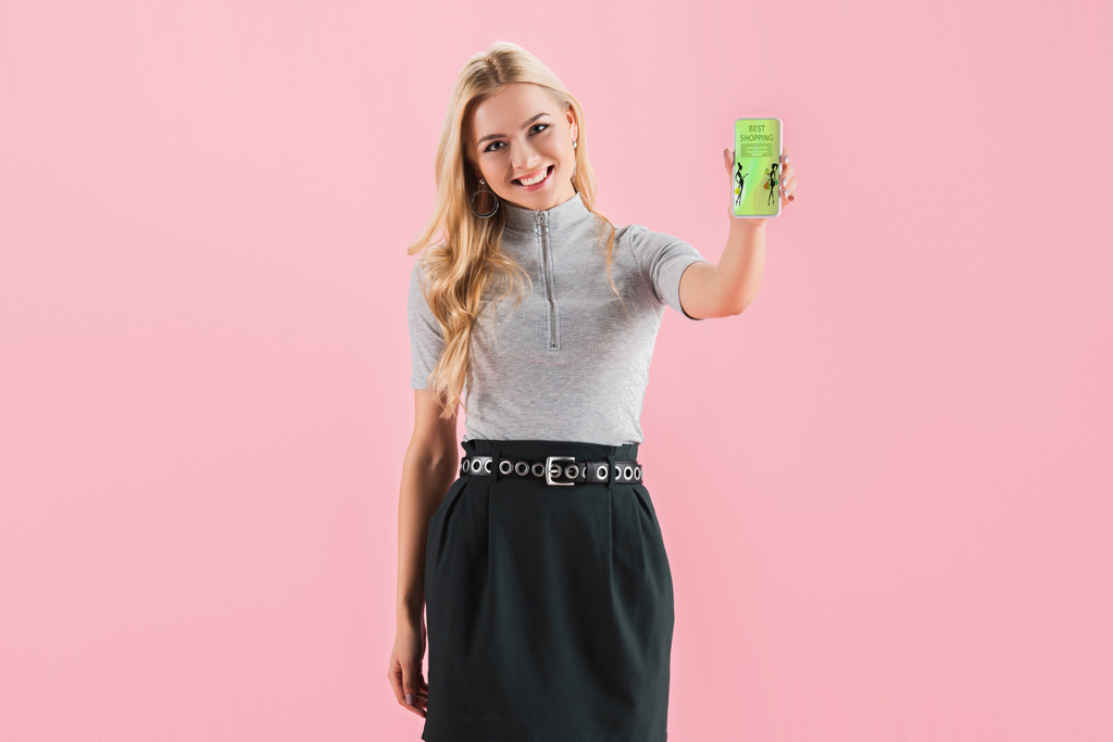улыбающаяся блондинка, показывающая смартфон с приложением для покупок на экране, изолированная на розовом
 - Фото, изображение