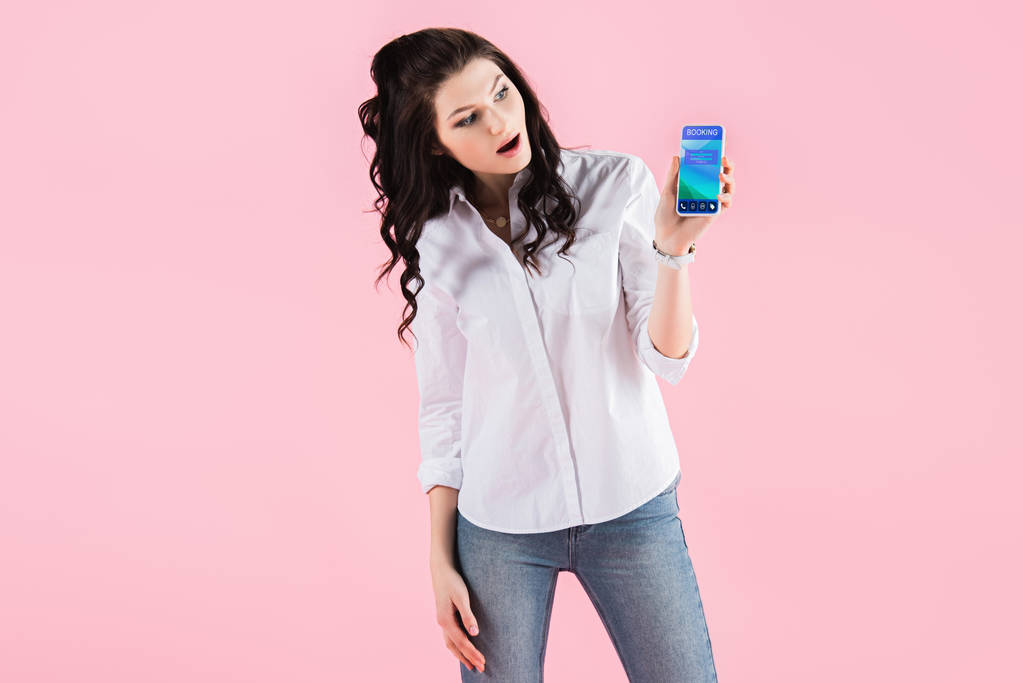 šokovaná žena ukazuje smartphone s rezervační aplikace na obrazovce, izolované na růžové - Fotografie, Obrázek
