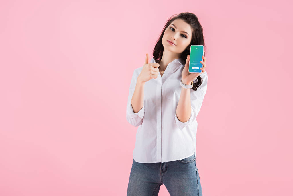 привлекательная брюнетка показывает большой палец вверх и смартфон с приложением Twitter на экране, изолированные на розовый
  - Фото, изображение