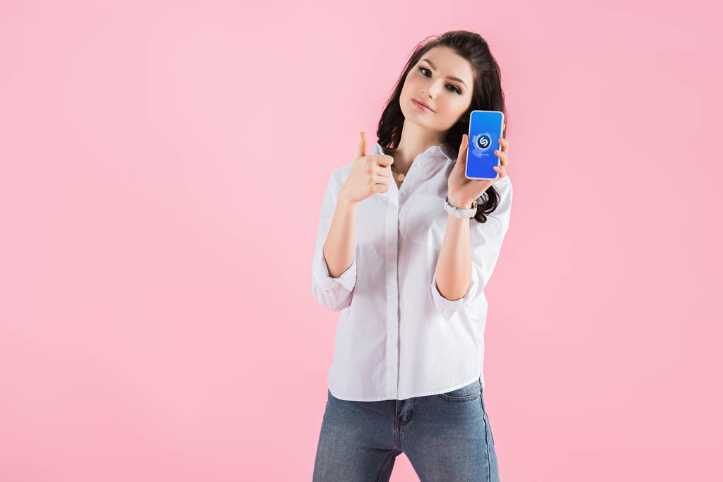 attraktive brünette Mädchen zeigt Daumen hoch und Smartphone mit Shazam-App auf dem Bildschirm, isoliert auf rosa  - Foto, Bild
