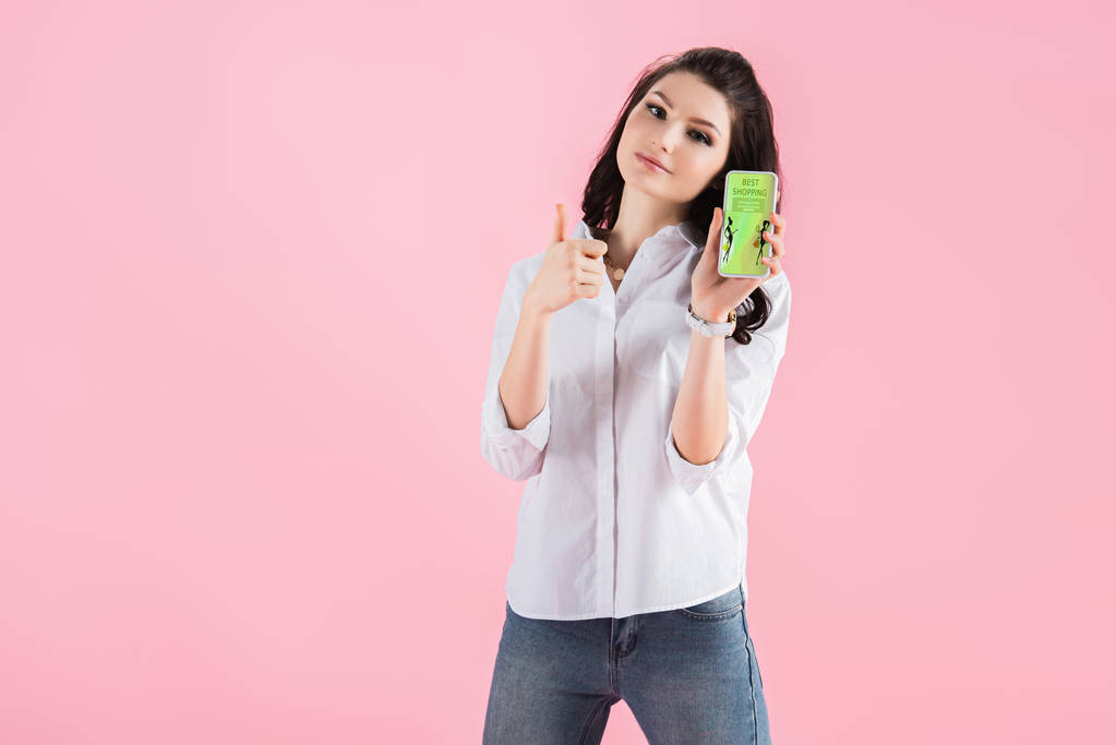 красивая девушка показывает большой палец вверх и смартфон с приложением для покупок на экране, изолированный на розовом
 - Фото, изображение