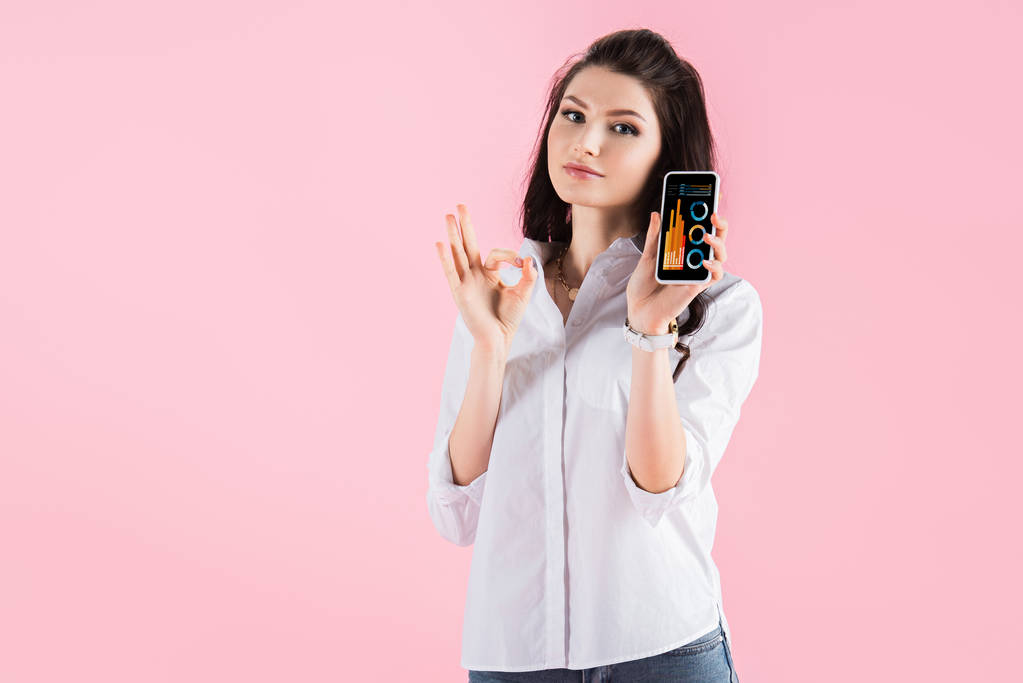 ελκυστικές κορίτσι μελαχρινή δείχνει εντάξει σημάδι και smartphone με γράφημα app στην οθόνη, απομονώνονται σε ροζ - Φωτογραφία, εικόνα