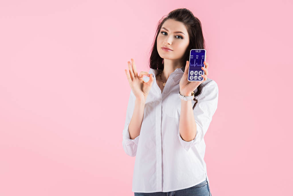 attraente ragazza bruna mostrando segno ok e smartphone con dati di salute sullo schermo, isolato su rosa
 - Foto, immagini