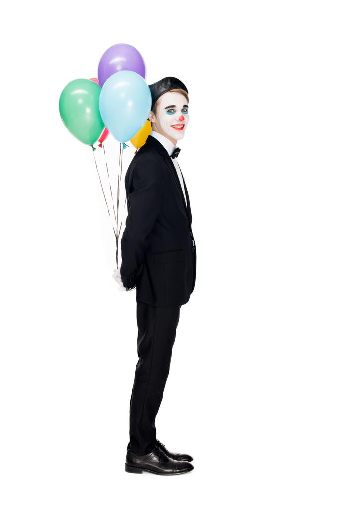 payaso alegre en traje y boina negra sosteniendo globos de helio detrás de la espalda aislados en blanco
  - Foto, imagen