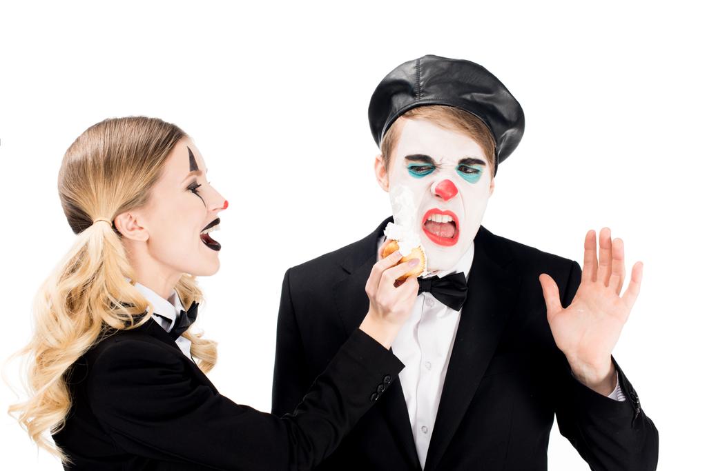 весёлая женщина-клоун бросает кекс в лицо недовольного мужчины, изолированного на белом
  - Фото, изображение