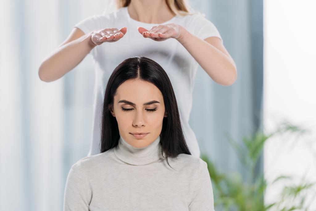 cropped shot of young woman with closed eyes sitting and receiving reiki healing therapy above head  - Photo, Image