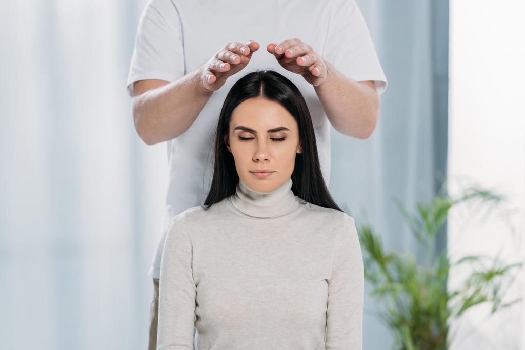 abgeschnittene Aufnahme einer ruhigen jungen Frau mit geschlossenen Augen, die eine Reiki-Behandlung über dem Kopf erhält  - Foto, Bild