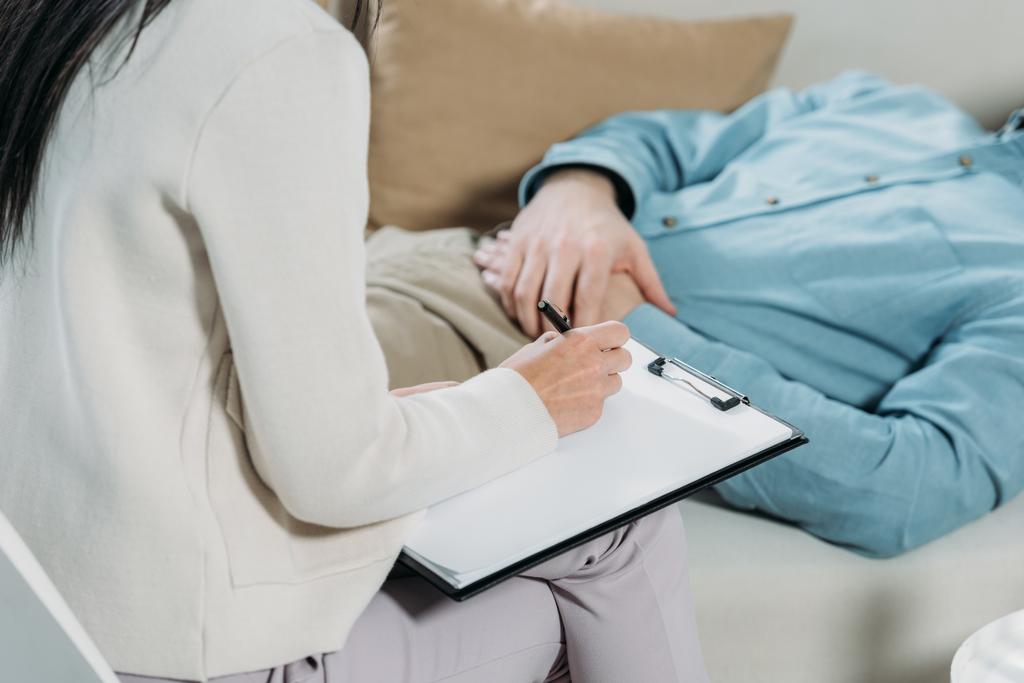 クリップボードとソファに横たわっている男性患者を書く精神療法医のクロップ撮影 - 写真・画像