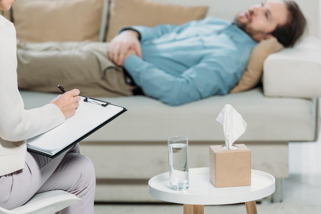 Schnappschuss eines Psychotherapeuten, der auf Klemmbrett schreibt, während ein bärtiger Mann auf der Couch liegt - Foto, Bild