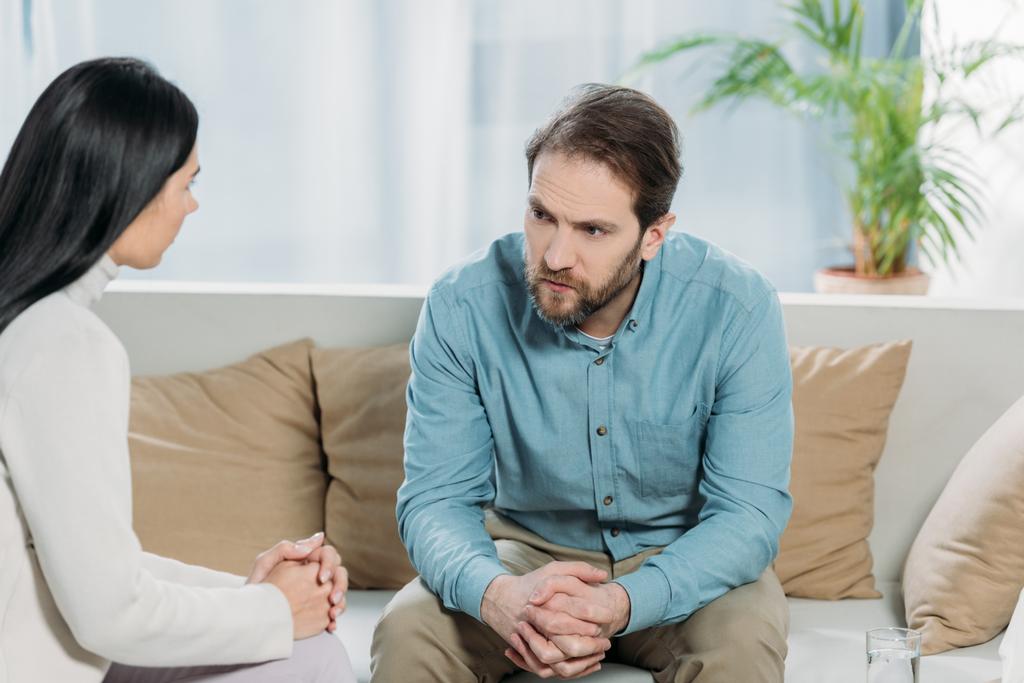 jonge vrouwelijke psychotherapeut en boos bebaarde man zitten en kijken naar elkaar - Foto, afbeelding