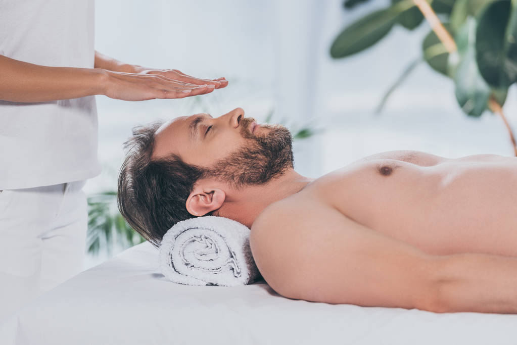 cropped shot of bearded man with closed eyes lying and receiving reiki therapy above head - Photo, Image