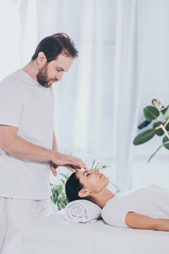 side view of bearded healer doing reiki treatment session to young woman with closed eyes - Photo, Image
