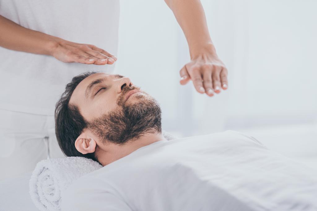 Ruhiger bärtiger Mann auf Massagetisch liegend und Hände des Heilers bei Reiki-Behandlung - Foto, Bild