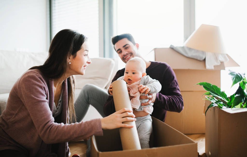 A portrait of happy young couple sitting on a sofa with a baby and cardboard boxes, moving in a new home. - Photo, Image