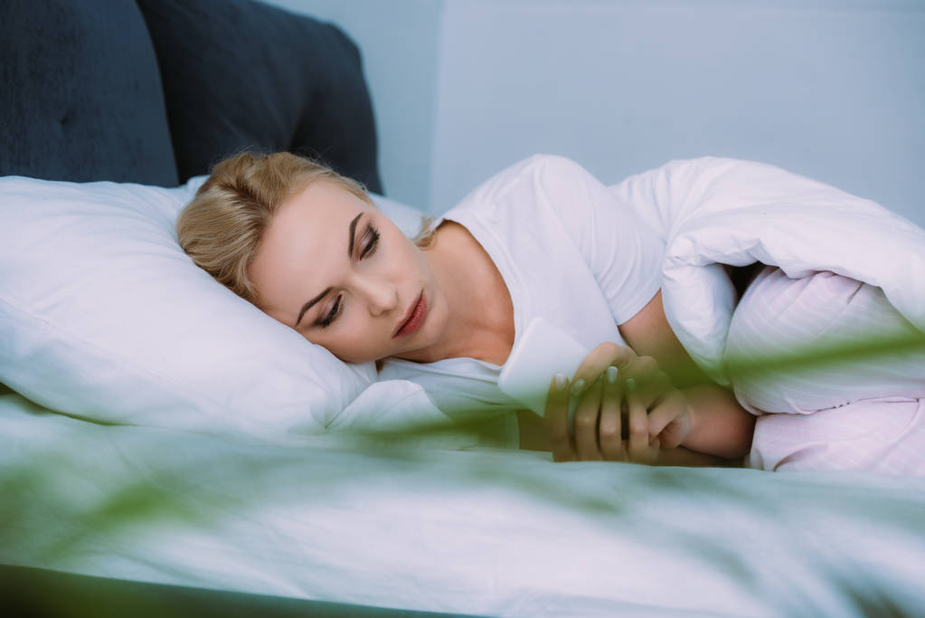 επιλεκτική εστίαση του όμορφη λυπημένος γυναίκα ξαπλωμένη στο κρεβάτι στο σπίτι και τη χρήση smartphone - Φωτογραφία, εικόνα