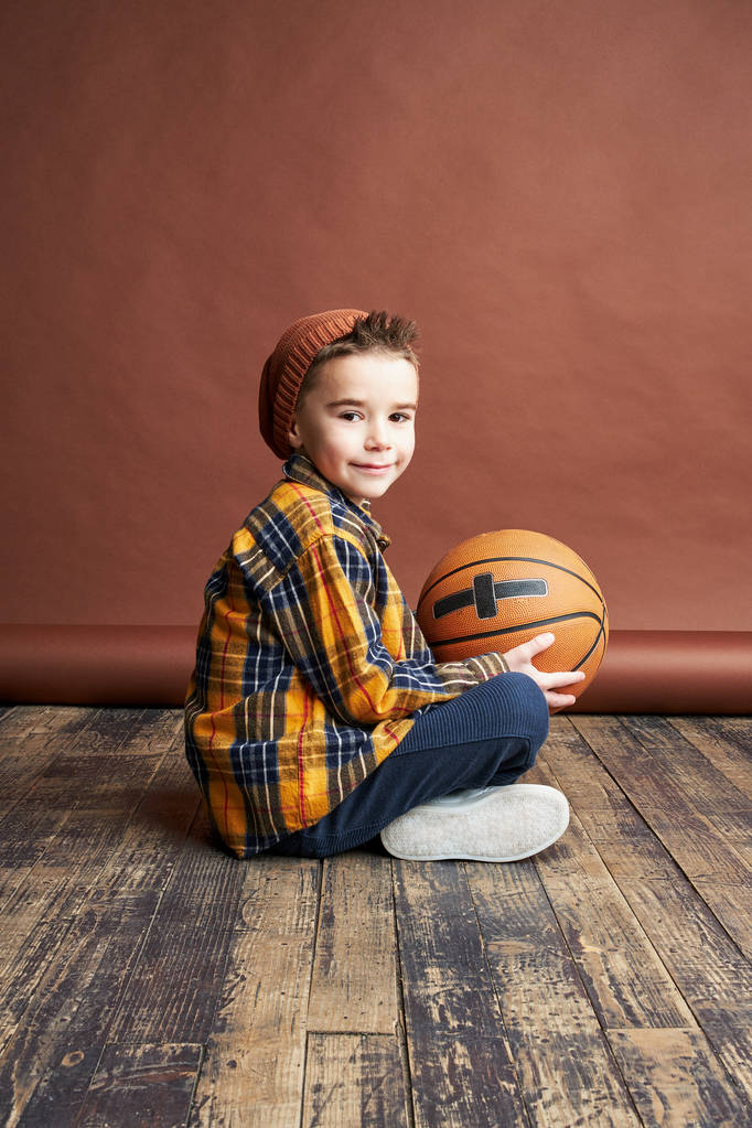おしゃれな男の子バスケット ボール ボールの手の中で床に座って。スタジオ撮影、茶色の背景  - 写真・画像