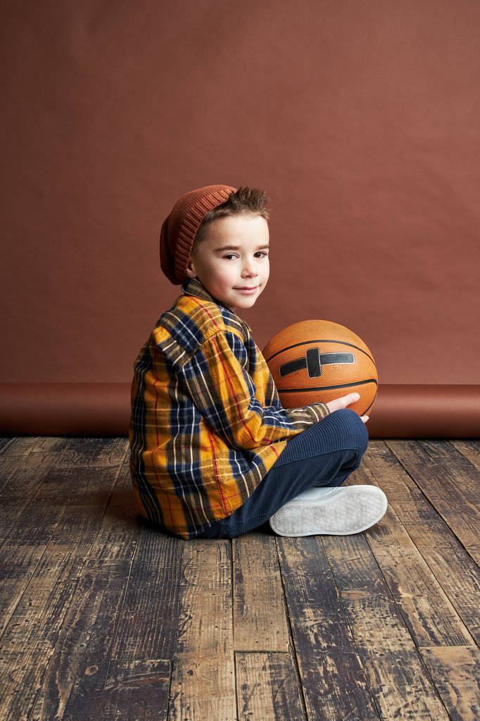 Κομψό αγόρι κάθεται στο πάτωμα με μπάλα του μπάσκετ στα χέρια. Studio που γυρίστηκε, καφέ φόντο  - Φωτογραφία, εικόνα