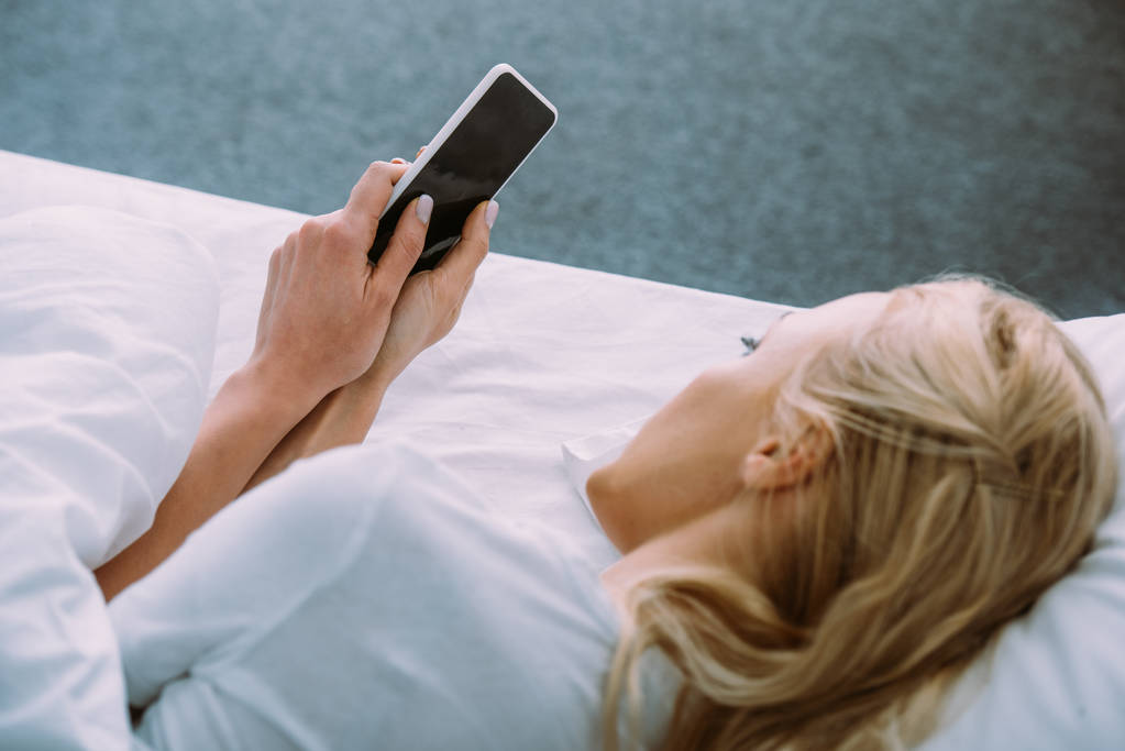 mujer solitaria acostada en la cama y usando un teléfono inteligente con pantalla en blanco
 - Foto, Imagen