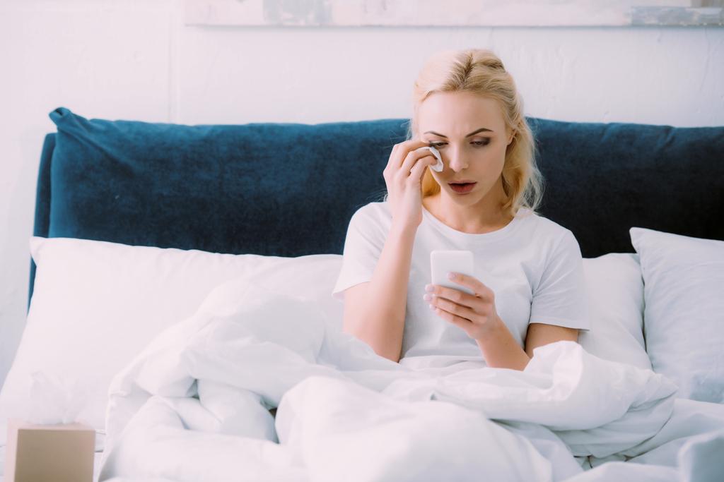κλάμα γυναίκα σκουπίζοντας δάκρυα και χρησιμοποιώντας το smartphone στο κρεβάτι στο σπίτι - Φωτογραφία, εικόνα