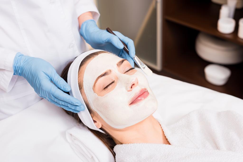 ayakta ve güzellik salonu yüzündeki kadın kozmetik maskesi yakınındaki kozmetik fırça tutan güzellik uzmanı - Fotoğraf, Görsel