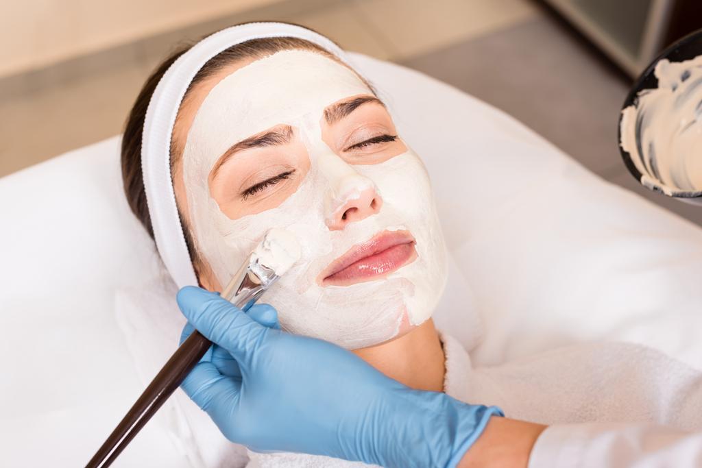 косметолог наносит косметическую маску на лицо женщины косметической щеткой в салоне красоты
 - Фото, изображение