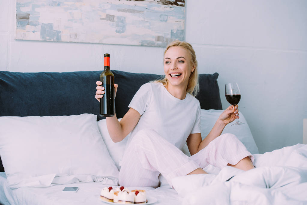 glückliche Frau mit Flasche und Glas Rotwein, während sie Geburtstag allein im Bett feiert - Foto, Bild